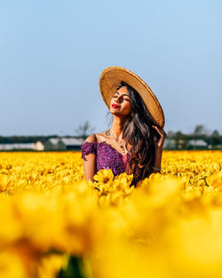 Woman wearing hat while standing by yellow flowering plants against clear sky