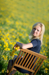 Portrait of young woman holding floral crown while sitting on chair at dandelion meadow