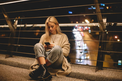 Teenage girl using smart phone sitting on bridge in city at night