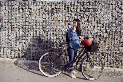 Full length of woman looking away while standing with bicycle against stone wall
