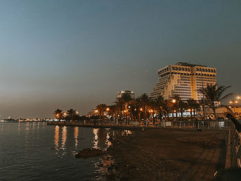 Illuminated modern buildings by sea against sky at night