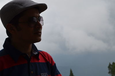 Man wearing sunglasses and cap while looking away against sky