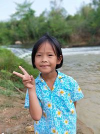 Portrait of cheerful cute girl showing peace symbol while standing on shore by river