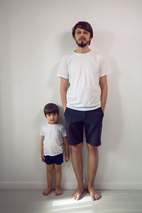 Father and son in white t-shirts and shorts stand at home against the wall