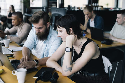 Male computer programmer coding on laptop by non-binary colleague in startup company