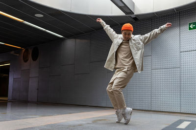 Full body happy young asian male wearing light casual outfit and orange hat standing with arms outstretched on tiptoes and looking down with smile