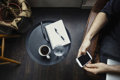 Cropped image of businessman using phone while sitting by drinks and spiral notebook