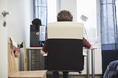 Rear view of teenage boy wearing headphones while sitting on chair at home