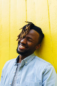 Young man with dreadlocks laughing in front of yellow wall