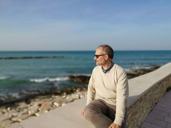 Man looking at sea while sitting on retaining wall against sky