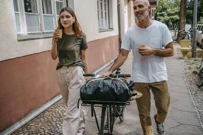 Male and female friends having coffee while wheeling with bicycle on sidewalk