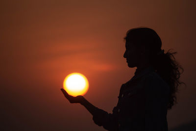 Optical illusion of silhouette woman holding sun standing against sky during sunset