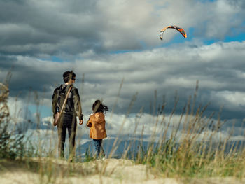 Back view of unrecognizable woman holding hand of daughter while walking together on hilly sandy seashore against wavy ocean and cloudy sky in windy weather