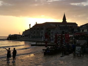 Silhouette people standing on sea by city against sky during sunset