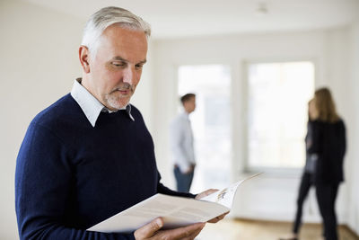 Mature real estate agent reading brochure with colleague and couple in background at home