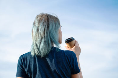 Unrecognisable blue haired woman drinks take away coffee against blue sky.