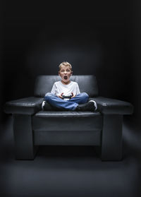 Portrait of boy playing video game on chair against black background