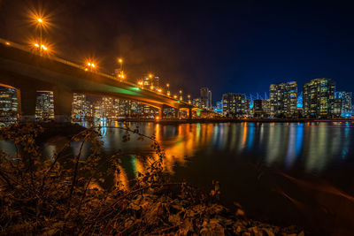 Illuminated bridge over river by buildings against sky at night