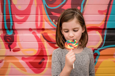 A young girls eats a huge rainbow lollipop in front of a colorful wall