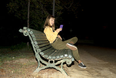 Full length of young woman using mobile phone sitting on bench at park