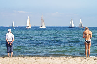 Rear view of young woman in bikini and man standing on shore at beach against sky