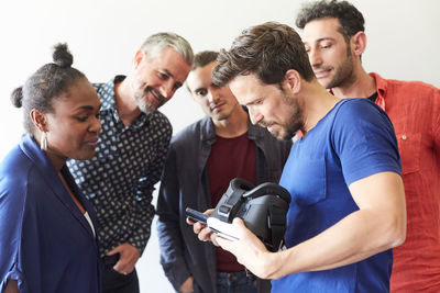 Businessman holding virtual reality simulator while showing mobile phone to colleagues in creative office