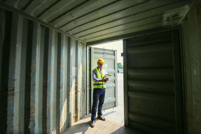 Full length of manual worker holding clipboard standing under cargo container