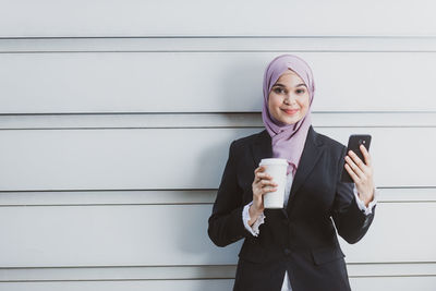 Portrait of businesswoman holding coffee while using mobile phone against wall