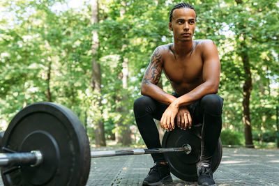 Full length of shirtless young man sitting on barbell outdoors