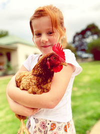 Portrait of smiling girl holding hen while standing in yard