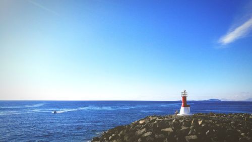 Lighthouse by sea against clear blue sky