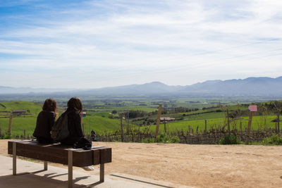 Female friends sitting on bench with landscape in background against sky