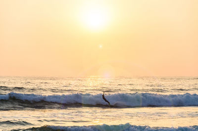 Silhouette man surfing in sea against bright sun in sky during sunset
