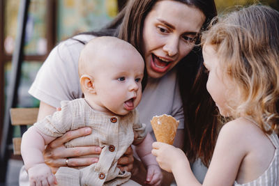 Caucasian toddler girl feeding baby brother boy with ice cream cone. mother is laughing. summer