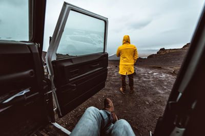 Low section of man sitting in car while friend looking at landscape against sky