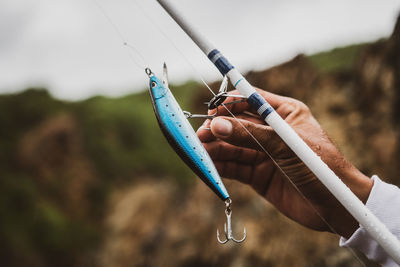 Crop anonymous male with special blue plug on fishing rod with hook standing in nature on blurred background during fishing