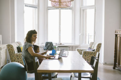 Mother using laptop while sitting with baby girl at table in house