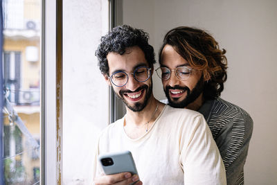 Man sharing smart phone with gay friend near window at home
