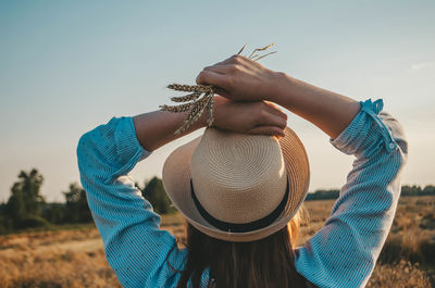 Rear view of woman wearing hat holding wheat against sky at sunset