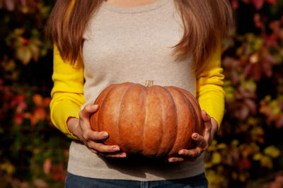 Close-up women hands holding large orange pumpkin on background autumn red leaves. colorful harvest 