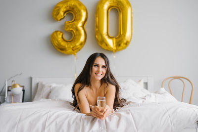 Happy young woman meets her thirtieth birthday in bed, holding a glass of champagne in her hands