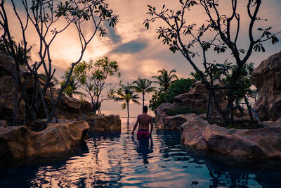 Man standing on rocks by swimming pool against sky during sunset