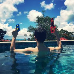 Boy holding toy gun while swimming in pool against sky