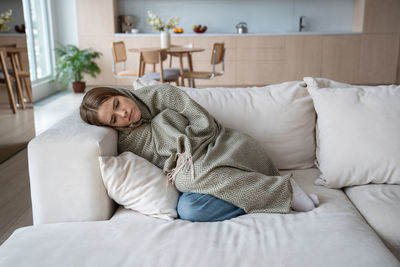 Unhealthy sad woman feeling cold covered with plaid lying on home sofa going through depression