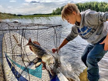 Full length of boy standing touching fish in net while standing by lake