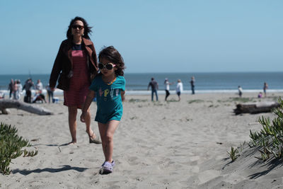 Mother and daughter walking at beach against clear sky on sunny day