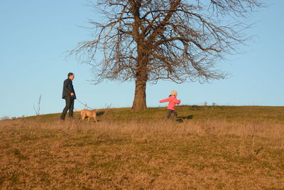 Father and daughter with dog walking on field against clear sky