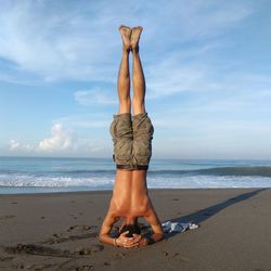 Rear view of shirtless young man doing headstand at beach against sky