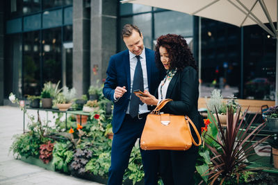 Smiling businessman and businesswoman sharing mobile phone against building in city