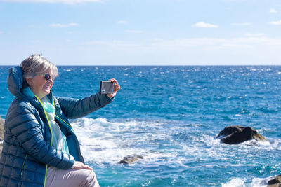 Smiling senior woman photographing by sea against sky